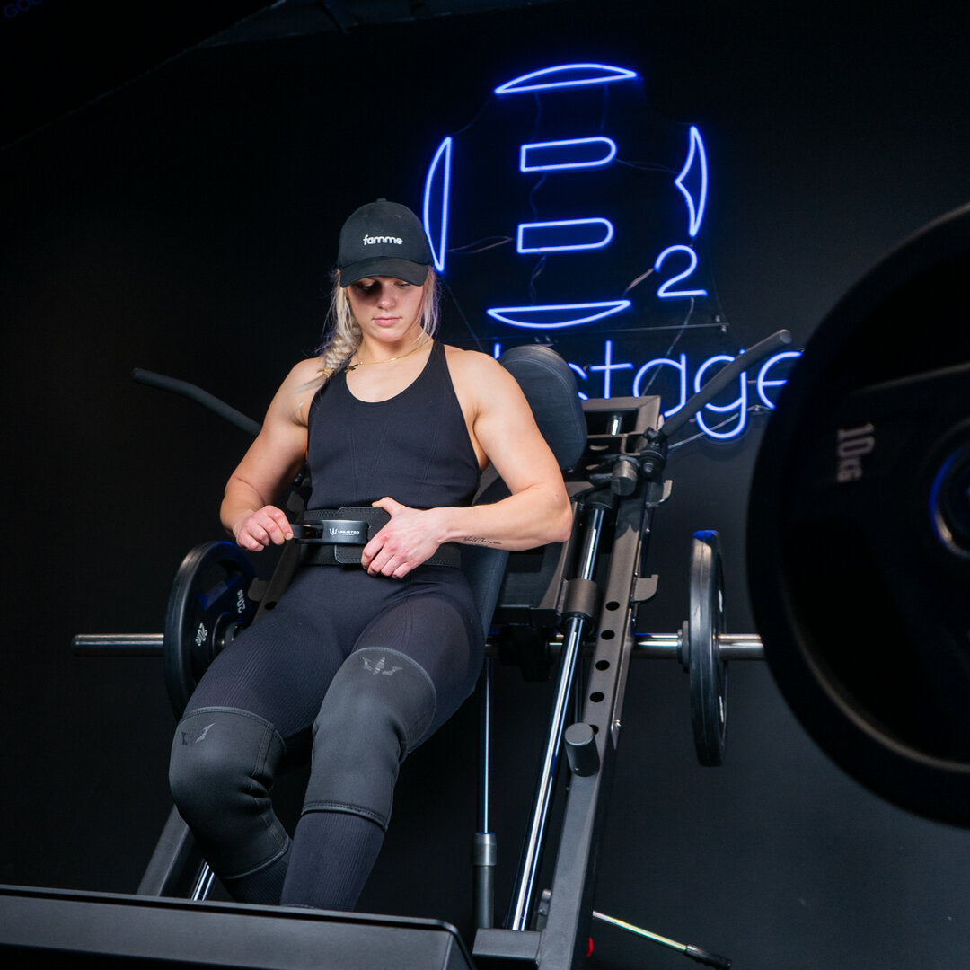 kvinde sidder i hacksquat maskine iført knee sleeves og er ved at lukke et lever belt foran et stort blåt neonskilt