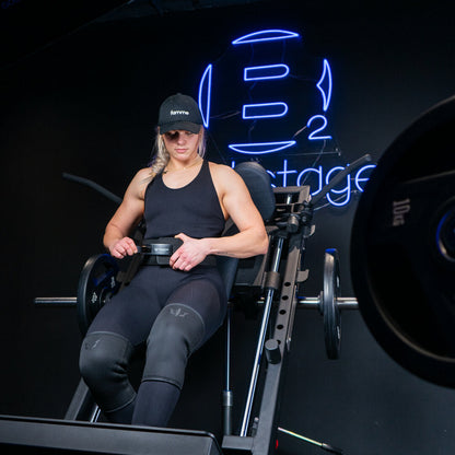 kvinde sidder i hacksquat maskine iført knee sleeves og er ved at lukke et lever belt foran et stort blåt neonskilt