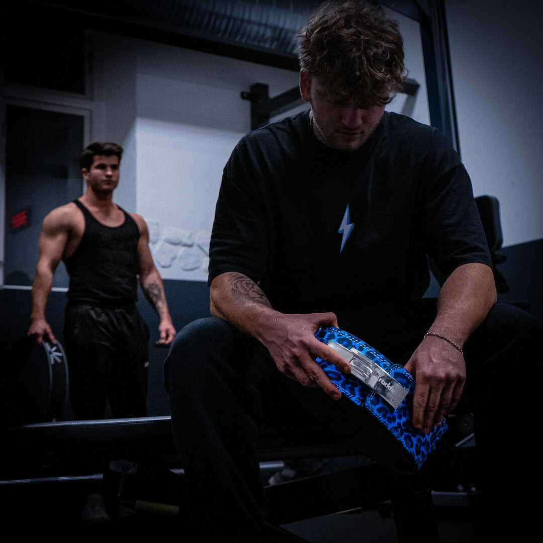 En mand sidder på bænk i træningscenter mens han holder et blåt Trackfie lever belt og kigger ned i gulvet, i baggrunden står en person med et andet Trackfie træningsbælte