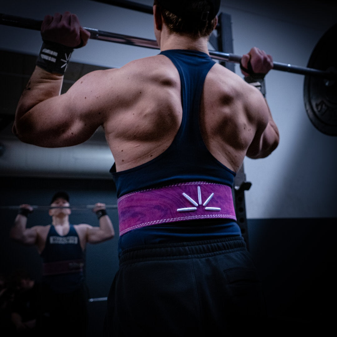 En mand shoulder presser og bruger wrist wraps og lilla træningsbælte i et fitnesscenter