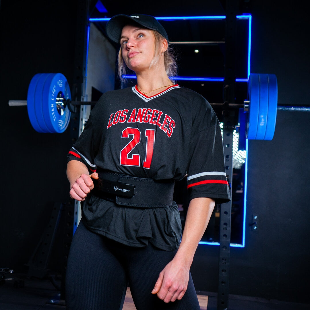 Kvinde står i fitnesscenter foran en squat rack iført et sort Unlisted Strength træningsbælte og kigger ud i luften