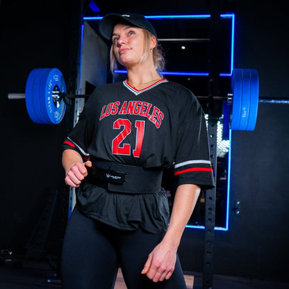 Kvinde står i fitnesscenter foran en squat rack iført et sort Unlisted Strength træningsbælte og kigger ud i luften